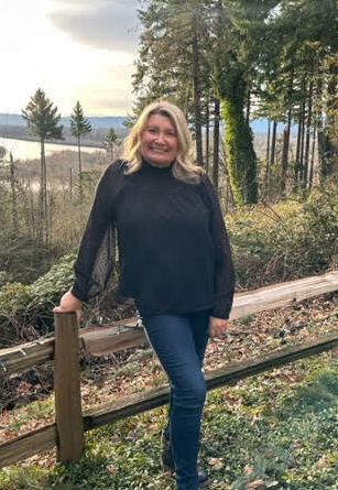 A photo of Carrie Schaller. She is in a grassy field leaning against a wooden rail. Behind her are trees and down the hill is lake