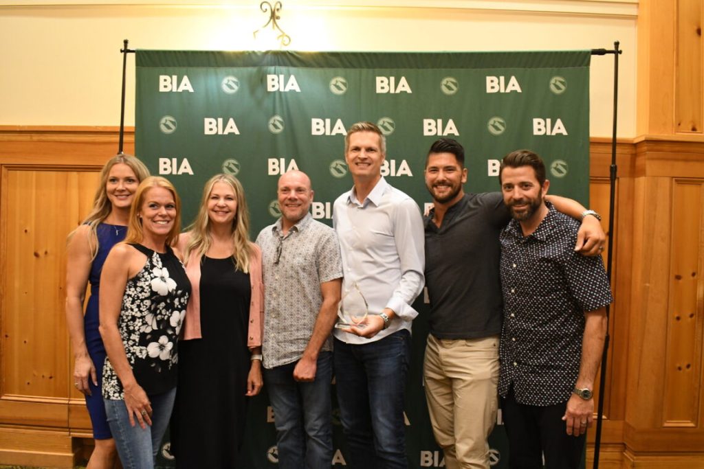 a group of 7 people are posing for a picture in front of a green background with the BIA logo printed on it in a repeating pattern. Individuals identified in the photo from Generation Homes Northwest are: Mandie, Ashley, Lester, Aaron, and Max